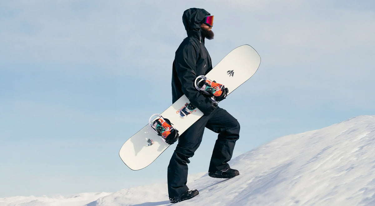 Snowboarder carrying a Jones snowboard up a snowy ridge